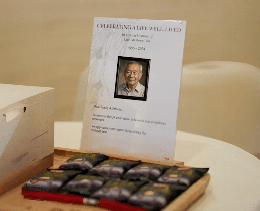 Memorial display at a Singapore Christian funeral service featuring a tribute card with a photo, condolence message, and snacks.